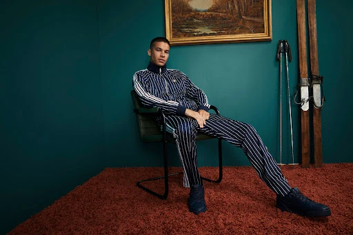 Man seated on a dark chair wearing a navy blue striped tracksuit against a teal wall with vintage skis.