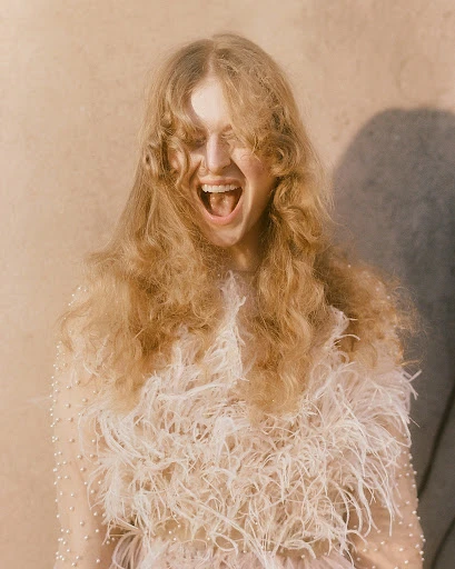 Portrait of a blonde woman with an open mouth wearing a feathered top standing indoors, photographed at eye-level against a neutral wall.