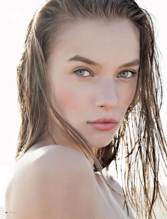 Close-up portrait of a woman with wet, slicked-back brown hair and natural makeup against a bright outdoor background