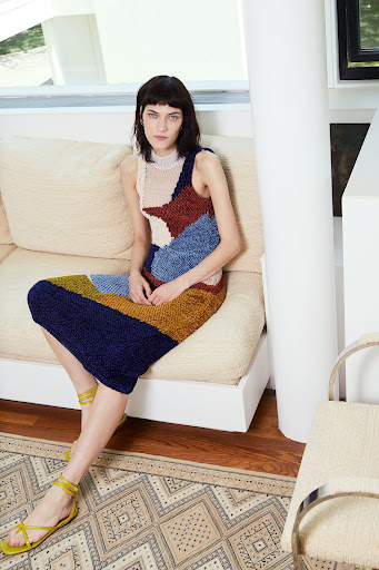 A woman is sitting on a couch wearing a multi-colored knit dress and sandals in an indoor setting with a patterned rug.