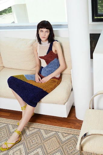 A woman is sitting on a couch wearing a multi-colored knit dress and sandals in an indoor setting with a patterned rug.