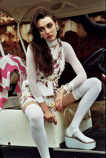 A woman with dark hair seated on a white vehicle wearing a floral dress, white stockings, and platform sandals.