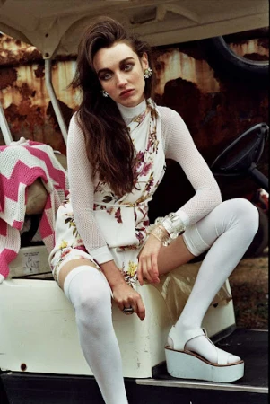 A woman with dark hair seated on a white vehicle wearing a floral dress, white stockings, and platform sandals.