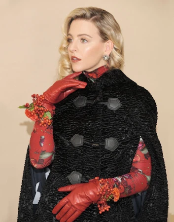 Blonde woman wearing a black textured cape and red leather gloves against a plain neutral background.