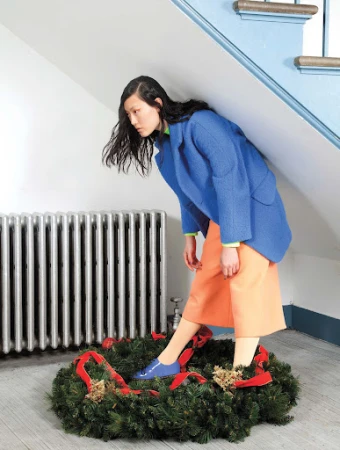 Fashion shot of a woman in a blue blazer and orange skirt standing inside of a green Christmas wreath with red ribbons indoors.