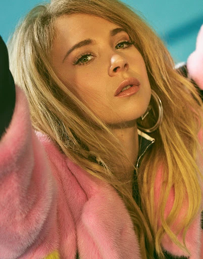 Close-up portrait of a blonde woman with hoop earrings and a pink fur coat, looking directly at the camera against a blue background.