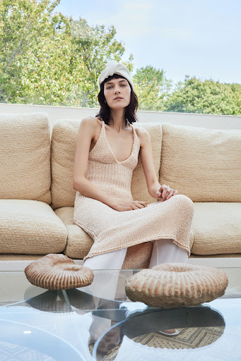 A woman in a knit dress sits on a couch in front of two decorative rock objects, with a trees and blue sky in the background.