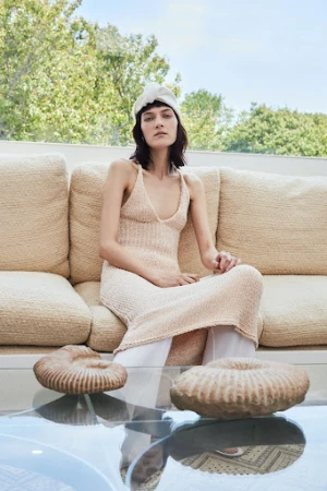 A woman in a knit dress sits on a couch in front of two decorative rock objects, with a trees and blue sky in the background.
