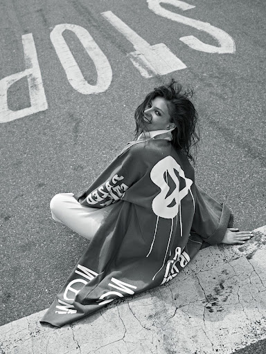 Woman sits on concrete next to street with large 'STOP' painted on asphalt. She wears a printed jacket, smiling. Black and white.