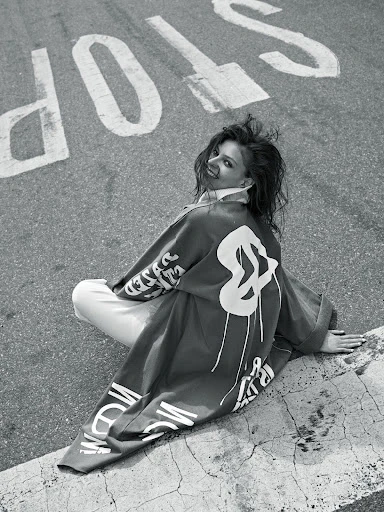 Woman sits on concrete next to street with large 'STOP' painted on asphalt. She wears a printed jacket, smiling. Black and white.