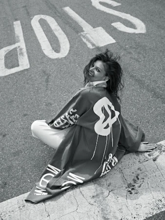 Woman sits on concrete next to street with large 'STOP' painted on asphalt. She wears a printed jacket, smiling. Black and white.