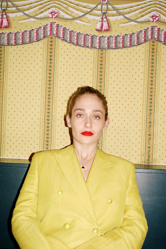 Portrait of a poised adult woman in a yellow blazer and red lipstick, gazing forward against a yellow patterned backdrop.