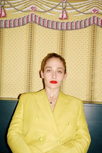 Portrait of a poised adult woman in a yellow blazer and red lipstick, gazing forward against a yellow patterned backdrop.