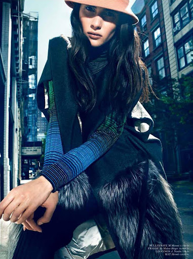 Model wearing a bucket hat, blue striped sleeves, and dark fur-trimmed outerwear on an urban street.