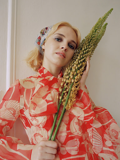 Woman in floral dress with flower. Portrait of a blond woman wearing a headscarf and holding a long flower against a white wall.