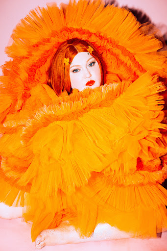 Red-haired adult female portrait, wearing a large orange ruffled dress, posing on a pink background with red lipstick and orange hair clips.