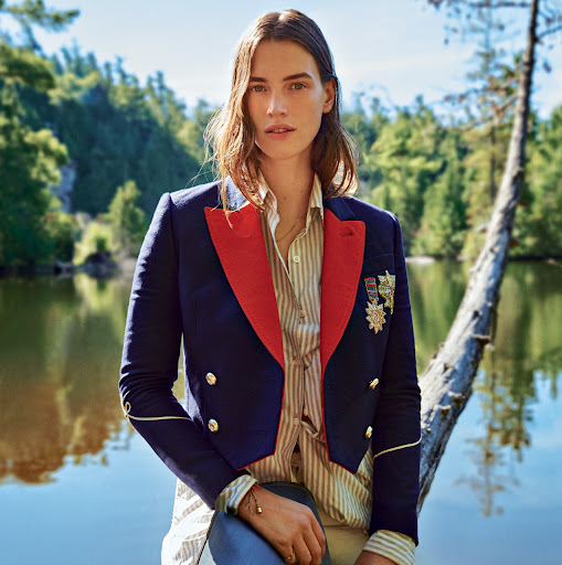 Woman in a navy and red military style blazer stands on a fallen log next to a lake surrounded by a green forest on a sunny day.