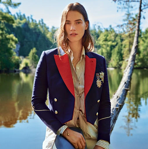 Woman in a navy and red military style blazer stands on a fallen log next to a lake surrounded by a green forest on a sunny day.