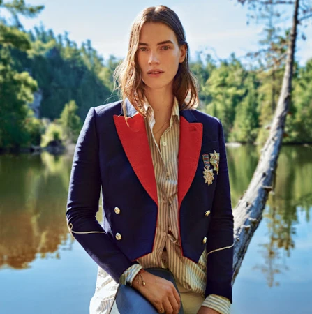 Woman in a navy and red military style blazer stands on a fallen log next to a lake surrounded by a green forest on a sunny day.