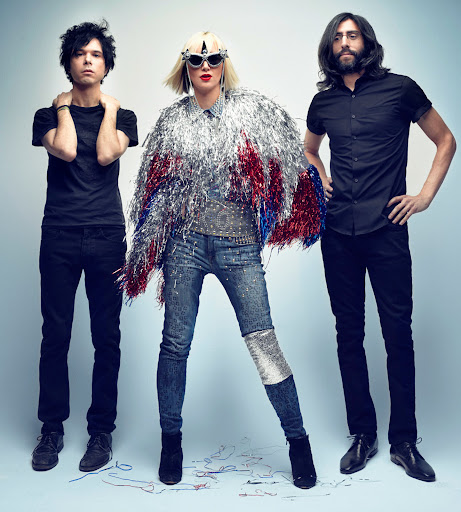 Three people in a photo studio, a woman wearing an elaborate reflective fringe jacket with two men standing on either side.