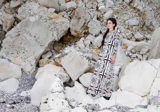 Adult female fashion model in a patterned maxi dress standing among massive white rocks, posing in a natural, rocky environment.