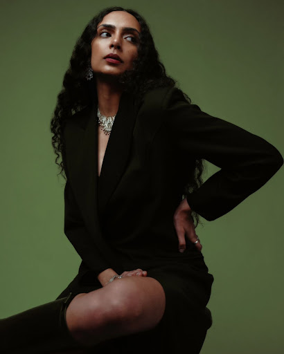 Portrait of a woman with long curly hair, wearing a black blazer and statement necklace, seated against a green studio backdrop.