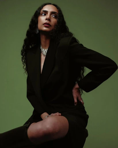 Portrait of a woman with long curly hair, wearing a black blazer and statement necklace, seated against a green studio backdrop.