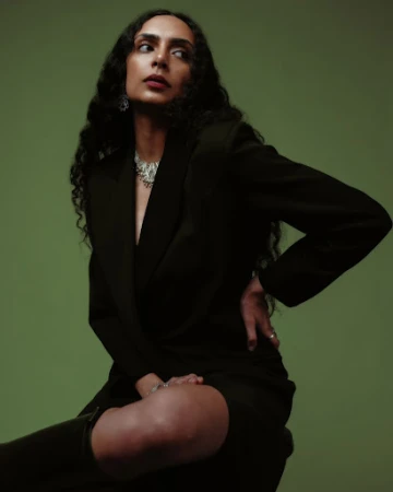 Portrait of a woman with long curly hair, wearing a black blazer and statement necklace, seated against a green studio backdrop.