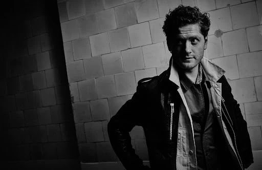 Monochrome portrait of a man in a dark leather jacket and striped shirt against a tiled wall, with grooming by Sheridan Ward.