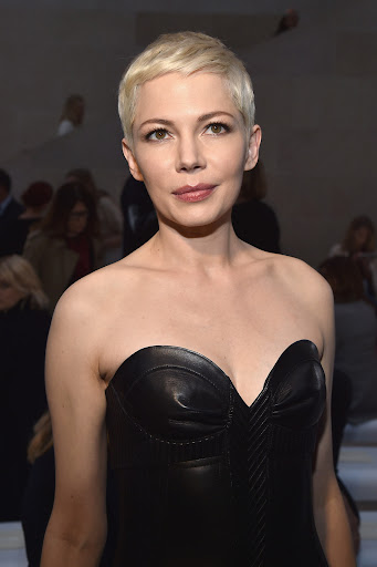 Close-up portrait of Michelle Williams with platinum blonde pixie cut wearing a black strapless dress with blurred background.