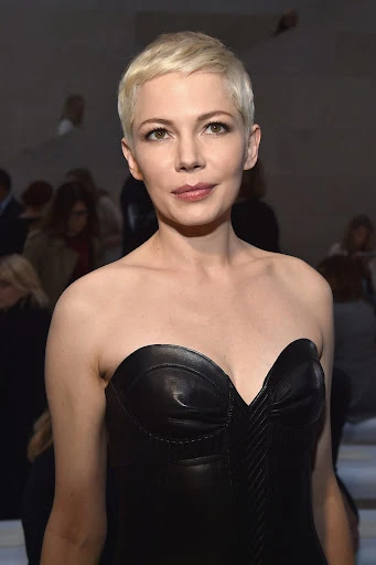 Close-up portrait of Michelle Williams with platinum blonde pixie cut wearing a black strapless dress with blurred background.