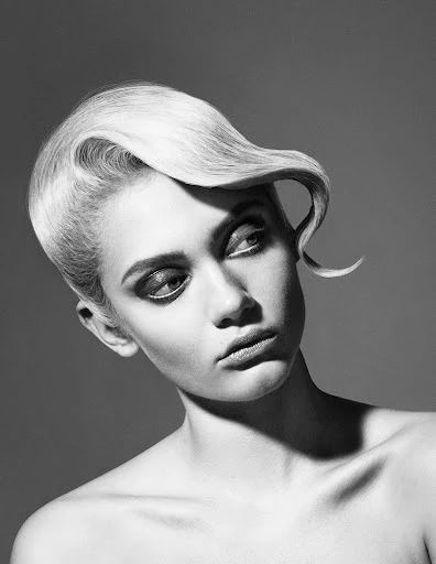 Monochrome portrait of a young adult woman with styled platinum blond hair looking down and to the left against a solid gray background.