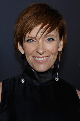 Close-up of Toni Collette smiling, wearing a black dress and long silver earrings against a dark background at an event.