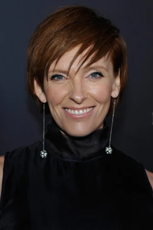 Close-up of Toni Collette smiling, wearing a black dress and long silver earrings against a dark background at an event.