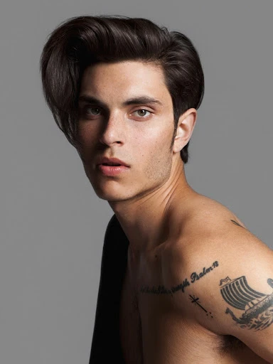Portrait of a young adult male model with styled dark hair, fair skin, and tattoos, posing indoors against a gray backdrop.