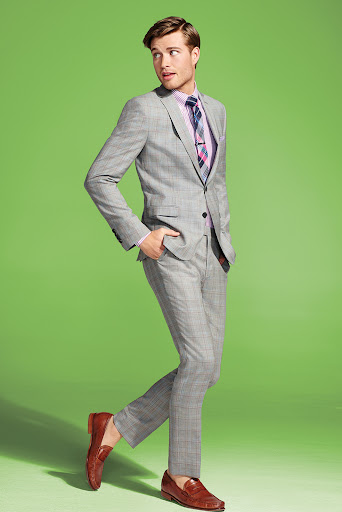 Man in gray plaid suit with pink shirt and tie posing against a lime green background. He is wearing brown loafers and looking to the left.