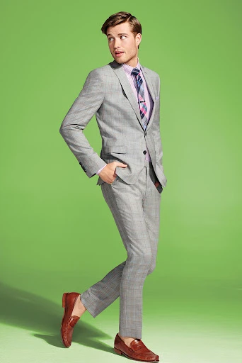 Man in gray plaid suit with pink shirt and tie posing against a lime green background. He is wearing brown loafers and looking to the left.