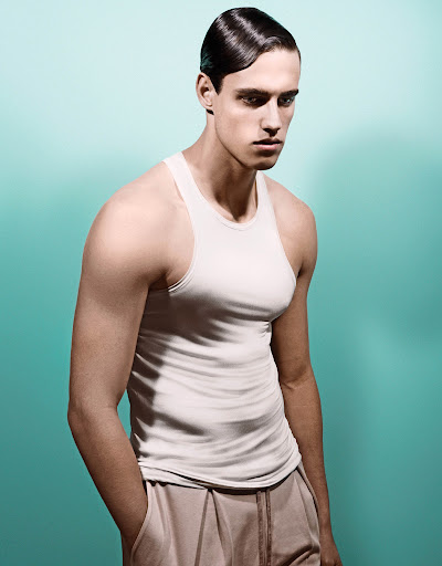 Portrait of a man with slicked hair wearing a white tank top against a cyan background, styled by Sheridan Ward.