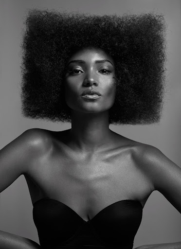Striking monochrome portrait of a dark-skinned woman with a large afro, wearing a strapless top, set against a neutral grey backdrop.