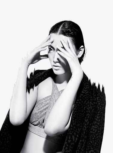 Striking black and white studio portrait of a woman with wet hair, gray crop top, and patterned jacket against a bright white background.