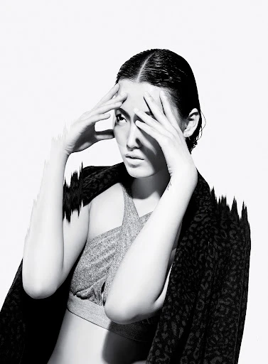 Striking black and white studio portrait of a woman with wet hair, gray crop top, and patterned jacket against a bright white background.