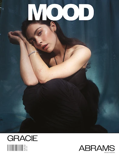 Gracie Abrams poses in a sleeveless black dress on a magazine cover with 'MOOD' as the title. The image has a teal background.