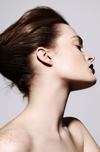 Portrait of a beautiful young woman with dark hair styled up, wearing dark lipstick, against a neutral grey background in a beauty editorial.
