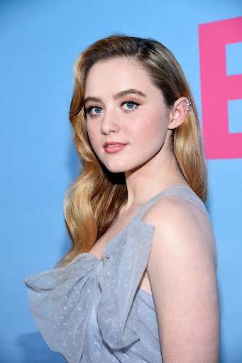 Portrait of actress Kathryn Newton with blonde wavy hair and a grey tulle dress against a blue backdrop