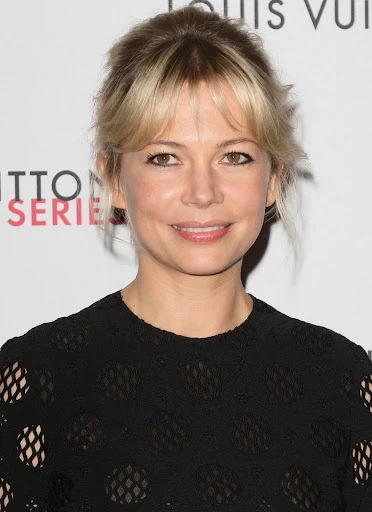 Close-up portrait of an adult woman with blonde hair and fair skin, wearing a black textured dress, with a fashion event backdrop.