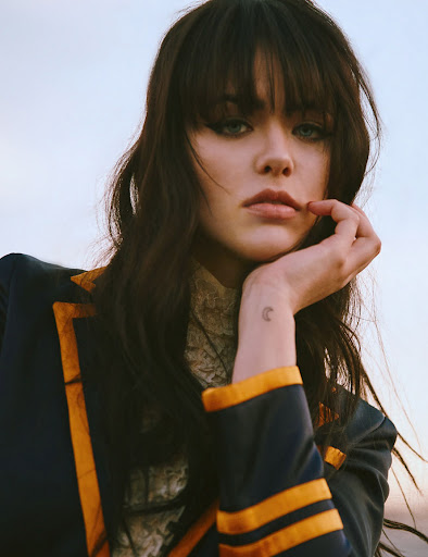 Portrait of a woman with dark hair wearing a navy and gold jacket, her hand resting on her face against a neutral sky background.