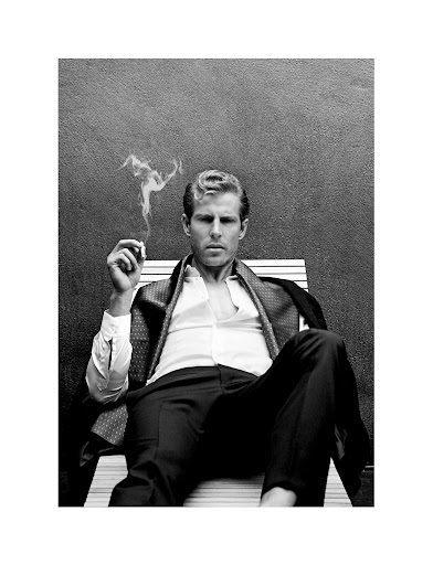 Black and white shot of a light-skinned adult man relaxing on a chair smoking a cigarette, wearing a jacket and dress shirt.