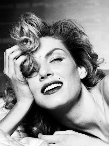 Black and white portrait of a smiling woman with curly hair, mole on her face, hand on her head, and laying on a bed.
