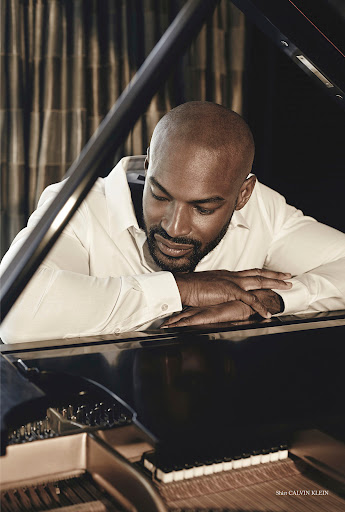 An adult male poses indoors with a grand piano in a portrait. The man is wearing a white dress shirt with dark curtains behind him.