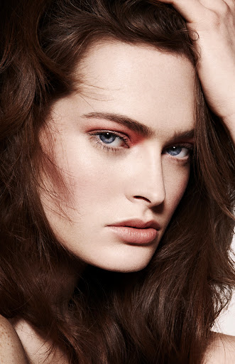Close-up beauty shot of a woman with auburn hair, fair skin, and bright blue eyes wearing pink eyeshadow and natural-toned lipstick in a photo studio.